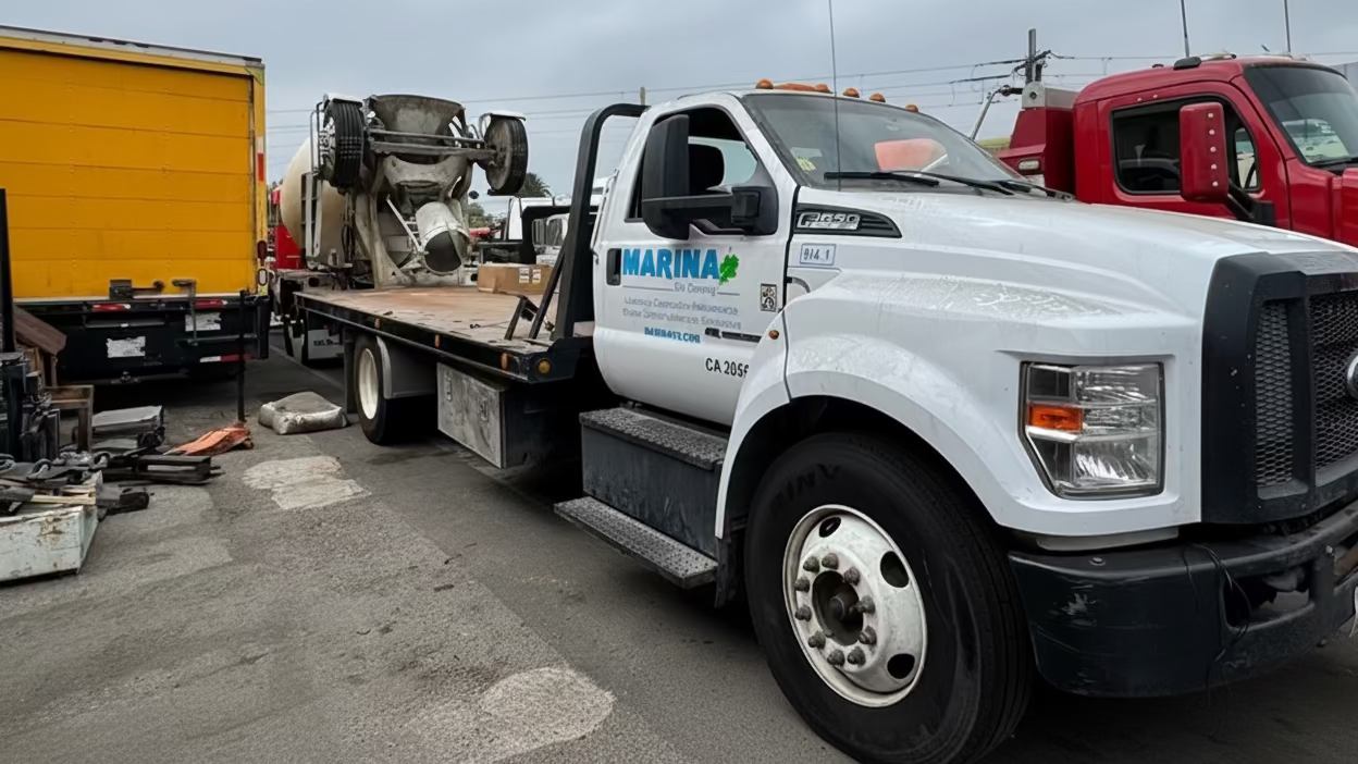 A white Ford flatbed tow truck with 'MARIAN' and 'CA 9597' printed on the side, parked in an outdoor lot alongside other trucks under a cloudy sky.
